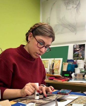 A woman is seated at a table, surrounded by various office supplies and a book. The indoor setting suggests an environment conducive to learning or work, with art on the walls enhancing the atmosphere.