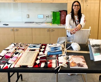 A woman is sitting in front of a table that has various items on it. The setting is indoors, featuring furniture like a desk and possibly a wall with art or design elements. The focus is on the woman and her attire, as well as the items displayed on the table.