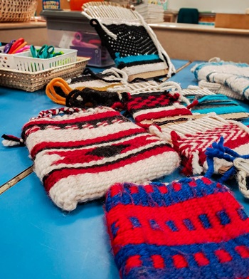 A group of colorful woven objects is displayed on a table. The scene showcases various textiles, highlighting the artistry of knitting and crochet. The vibrant threads and fibers indicate a creative crafting process, filled with intricate stitches and designs.