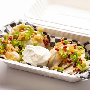 A portion of pierogi is served in a take-out container, garnished with bacon and green onions, and a side of sour cream.  The image features a close-up, eye-level shot of a serving of pierogi presented in a white, disposable take-out container. 