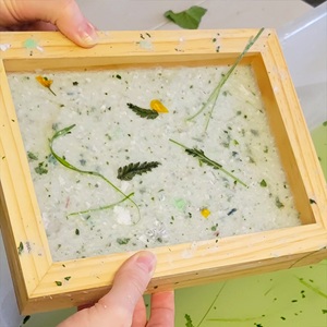 The image shows a close-up, high-angle view of a person's hands holding a wooden deckle and mold used for papermaking. The frame is made of light-colored wood, with a slightly rough texture, and appears to be handmade. Inside the frame, a sheet of wet, handmade paper pulp is visible. The pulp is a creamy white color and appears to be infused with various natural elements. These inclusions consist of small, fragmented green leaves, delicate blades of grass, and a few tiny yellow flower petals. 