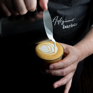 The image captures a close-up, overhead shot of a barista in the process of creating latte art. The scene is focused on the creation of a leaf or fern-like pattern in a cup of coffee.