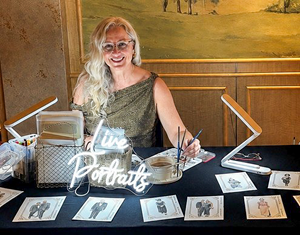 The image depicts a woman artist at her workspace, set up for creating live portraits. She is a middle-aged woman with long, wavy blonde hair and glasses, smiling warmly at the viewer. She is wearing a sleeveless, olive-green dress with a patterned texture. Her right hand is raised, holding a paintbrush, and she appears to be in the process of working or preparing to work.