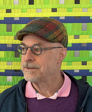 A medium shot portrait of a light-skinned, bald man with a mustache and goatee is captured against a vibrant, geometric background. He is looking to his left, with his head slightly tilted. The man is wearing a plaid flat cap in shades of brown, red, and green. He has round, dark-rimmed glasses on. His attire includes a bright pink polo shirt peeking out from under a deep purple knitted sweater.