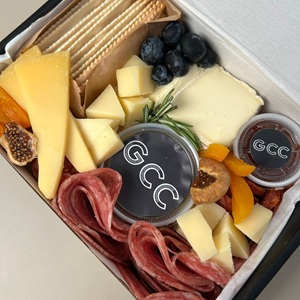 The image displays a close-up, top-down view of a charcuterie box, presented in a white cardboard container with dark gray accents. The box is densely packed with a variety of food items.