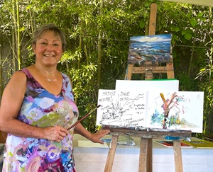 The image depicts a woman, identified as Jane Appleby, a visual artist, engaging in an art demonstration outdoors. She is positioned to the left of the frame, facing the viewer with a warm smile. She has short, graying hair, and is wearing a sleeveless dress with a vibrant, abstract floral pattern featuring hues of purple, pink, blue, and white. A delicate silver necklace adorns her neck. She is holding a paintbrush in her right hand, pointing it towards an easel that stands to her right.