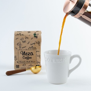 The image displays a close-up, eye-level shot on a clean white background, showcasing the process of pouring coffee. The main focus is on a white ceramic mug with the word "COFFEE" embossed on its side. A stream of dark brown coffee is being poured from a copper and glass French press, which is angled from the top right corner of the frame, with its lid slightly ajar and plunger mechanism visible. The coffee stream is a rich, dark amber color. 