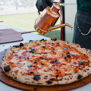 A close-up, eye-level shot captures a chef drizzling olive oil onto a freshly baked pizza. The pizza is the central focus, resting on a wooden peel, its crust puffed and speckled with dark, charred blisters, indicative of high-heat cooking. The topping is a vibrant combination of red tomato sauce and melted white mozzarella cheese, interspersed with fresh green basil leaves.