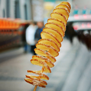 The image features a close-up, vertical shot of a spiral-cut potato, skewered and standing upright. The potato slices are thin and circular, arranged in a helix around a wooden skewer. Each slice is golden brown and appears to be lightly seasoned, with a dusting of white seasoning, possibly salt or sugar, visible along the edges. The texture of the potato looks crispy and slightly oily.