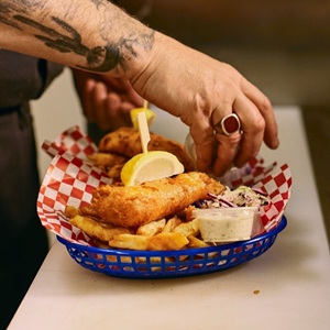 The image is a close-up, eye-level shot of a plate of fish and chips being prepared or served. The main focus is a blue plastic basket lined with red and white checkered paper. Inside the basket, there are several pieces of golden-brown, battered fish, and a generous portion of thick-cut french fries. A wedge of bright yellow lemon is perched on top of one of the fish pieces, secured with a wooden skewer. To the right of the fish and fries, a small clear plastic container holds a creamy white dipping sauce,