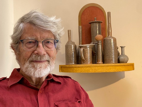 The image is a medium close-up portrait of Tam Irving with a display of pottery in the background. The man is positioned on the left side of the frame, facing slightly towards the viewer, and looking directly at the camera. He has a full head of gray hair, a thick gray beard, and is wearing glasses with dark frames. His expression is neutral and calm, with a slight hint of a smile. He is dressed in a red collared shirt with epaulets on the shoulders, suggesting a casual or work-oriented garment. The texture