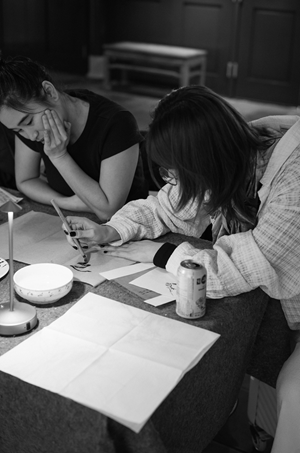 This is a black and white photograph capturing an intimate, focused moment between two women engaged in an artistic activity at a table. The image has a candid, documentary feel, emphasizing concentration and quiet interaction.