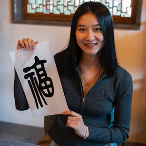 A woman with long, straight black hair is smiling warmly at the camera. She is wearing a dark gray, ribbed zip-up sweater and a delicate gold necklace with a small pendant. Her right hand holds up a white rectangular piece of paper displaying a large, bold black Chinese character: “福” (fú), which means “fortune” or “good luck.” Her left hand supports the bottom of the paper. The background features a traditional wooden window frame with intricate geometric lattice patterns, suggesting an East Asian architec