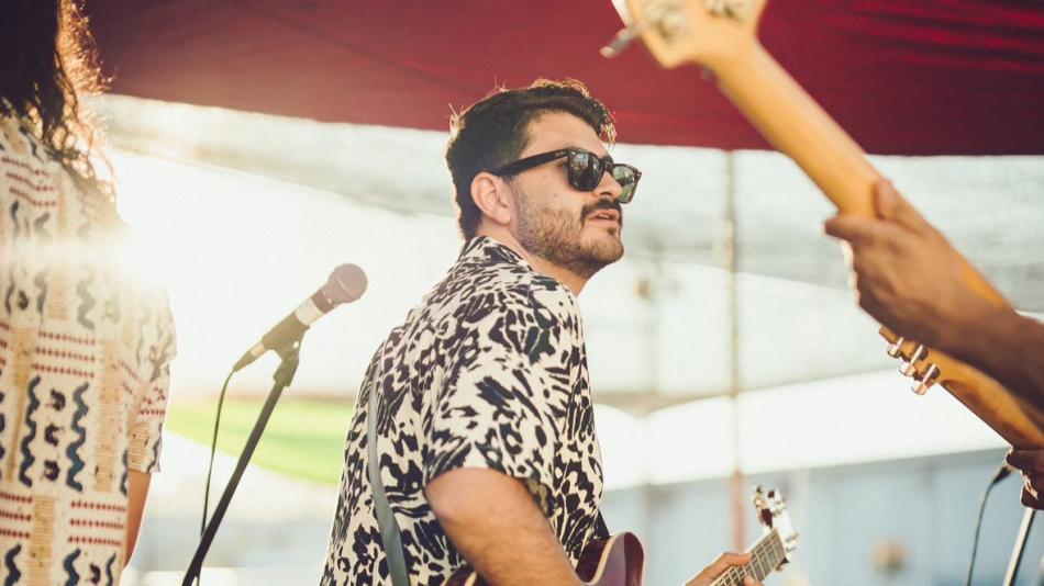 The main subject is a man in the mid-ground, seen from the chest up and in profile, looking to his right. He has dark hair, a beard, and is wearing dark-rimmed sunglasses. His shirt is the most striking detail of his attire, featuring a bold black and white pattern that resembles abstract animal prints or ink blots. He is holding a guitar, visible from the shoulder down, and a strap runs diagonally across his chest.