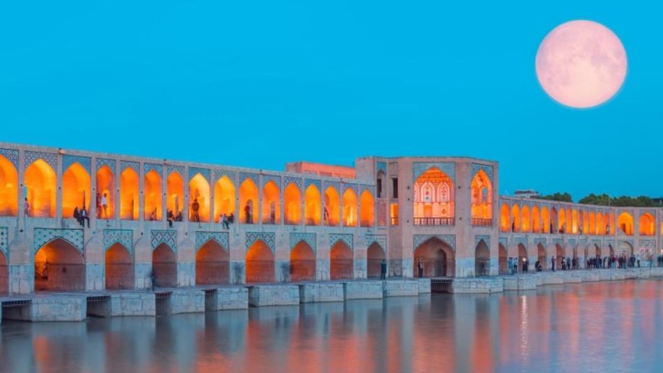 People can be seen resting in the Khaju Bridge in Iran. Key elements include the moon, water, building, sky, and reflection.
