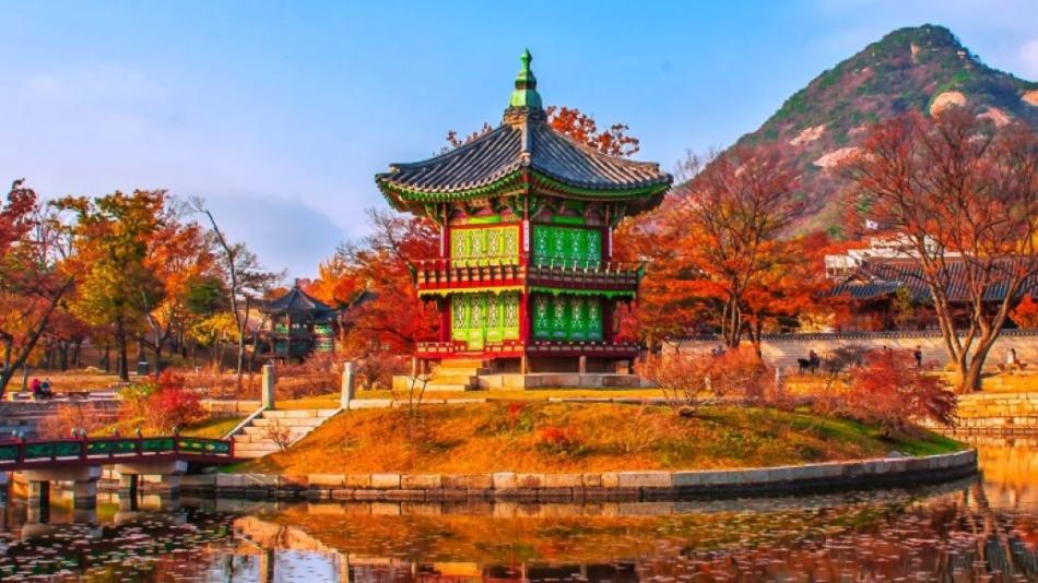 The photo depicts a building, possibly a temple or palace, with a green roof located by a body of water. In the background, there are trees and mountains. This scene is set in Korea.