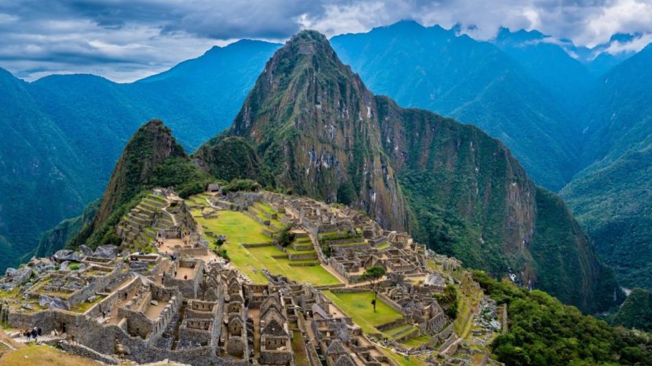 The image depicts a valley between mountains with Machu Picchu in the background. The setting is in Peru, showcasing a beautiful highland landscape with clouds, sky, and nature elements like grass and mountains.