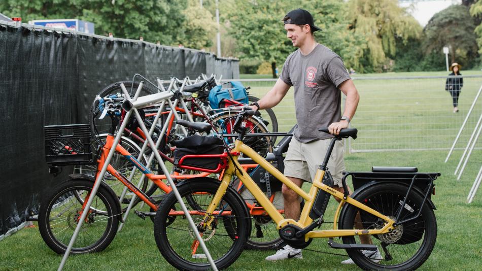 A man stands beside a bike, parking it at the bike valet in Ambleside Park, ready for a day of outdoor activities.