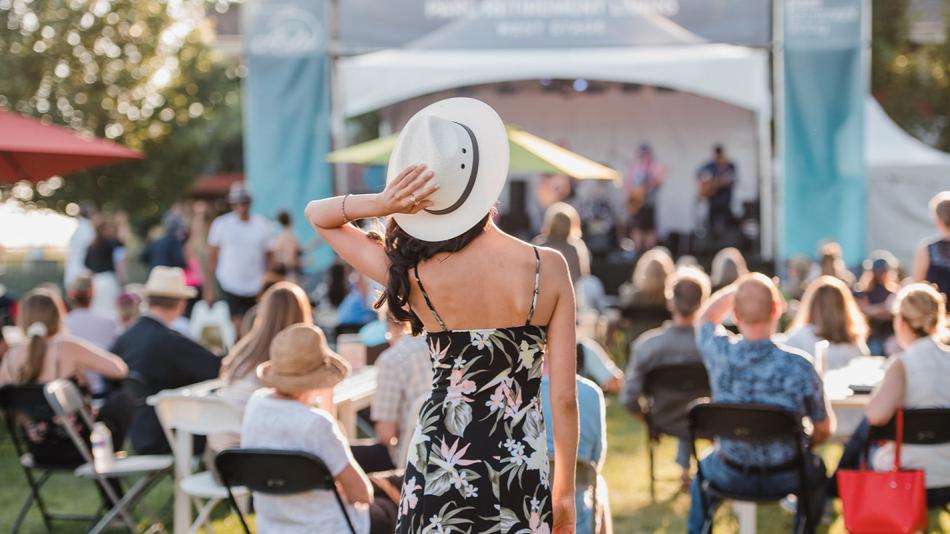 The image captures an outdoor event, likely a festival or concert, bathed in the warm glow of late afternoon or early evening sunlight. The scene is viewed from behind a woman in the foreground, who is the central subject. She has her back to the camera, wearing a black, spaghetti-strap dress with a bold floral pattern of pink and pale green flowers. She is also wearing a wide-brimmed white hat, tilted back slightly, with her right hand resting on its brim. A ring is visible on her right hand. Her dark hair