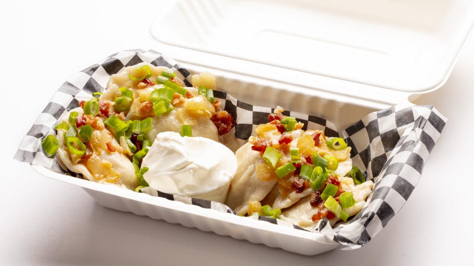 A portion of pierogi is served in a take-out container, garnished with bacon and green onions, and a side of sour cream.  The image features a close-up, eye-level shot of a serving of pierogi presented in a white, disposable take-out container. 