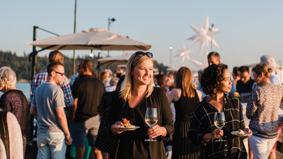 The image captures an outdoor social gathering at sunset, likely on a pier or boardwalk. In the foreground, two women are the focal points. The woman on the left, with blonde hair and sunglasses perched on her head, is smiling and looking to her right. She is holding a small white plate with a bit of food and a glass of white wine. She wears a black dress. Next to her, a woman with dark, curly hair is looking to her left, also holding a glass of white wine and a plate of food. She is wearing a black and whi