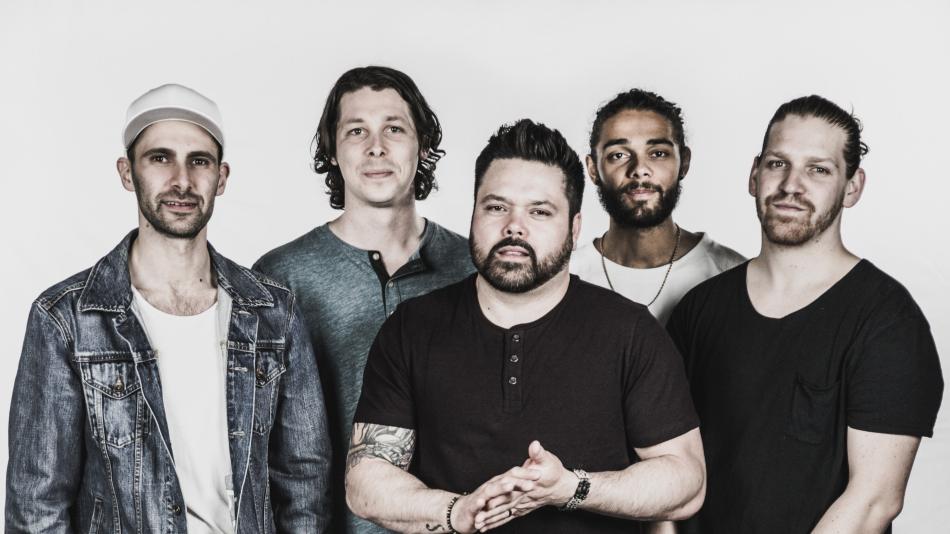 A group of five men stands in front of a plain white background, posing for a portrait. They are positioned in a slightly staggered line, with their torsos angled towards the camera. The lighting is soft and diffused, creating a gentle shadow on the left side of their faces. The overall color palette is muted, with a desaturated effect that lends a vintage or artistic feel to the image.