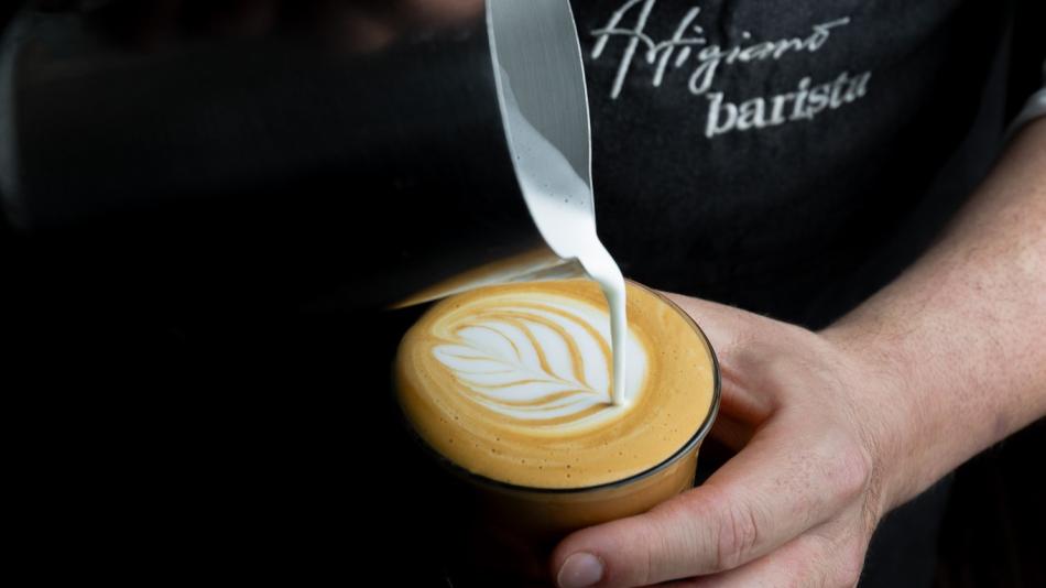 The image captures a close-up, overhead shot of a barista in the process of creating latte art. The scene is focused on the creation of a leaf or fern-like pattern in a cup of coffee.