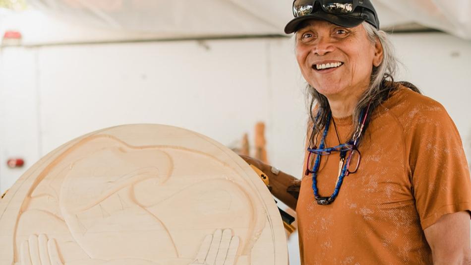 A man is holding a piece of wood indoors. He has a smile on his face and is wearing clothing that showcases his style. The setting includes an artistic wall, contributing to the overall ambiance of the scene.
