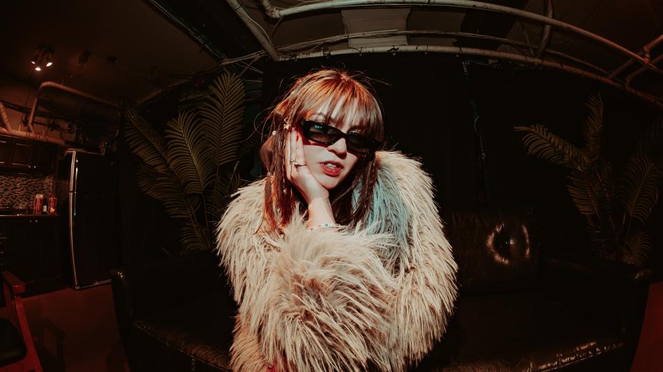 The image is a medium shot of a young woman posing in a dimly lit, somewhat cluttered indoor space. The woman is in the center of the frame, looking directly at the camera. She has reddish-brown hair with bangs and is wearing large, dark sunglasses with a thick black frame. Her left hand is resting on her cheek, with her fingers gently touching her face. She is wearing a light-colored, fluffy, faux fur jacket that covers most of her upper body. Several rings are visible on her left hand, and a beaded bracel