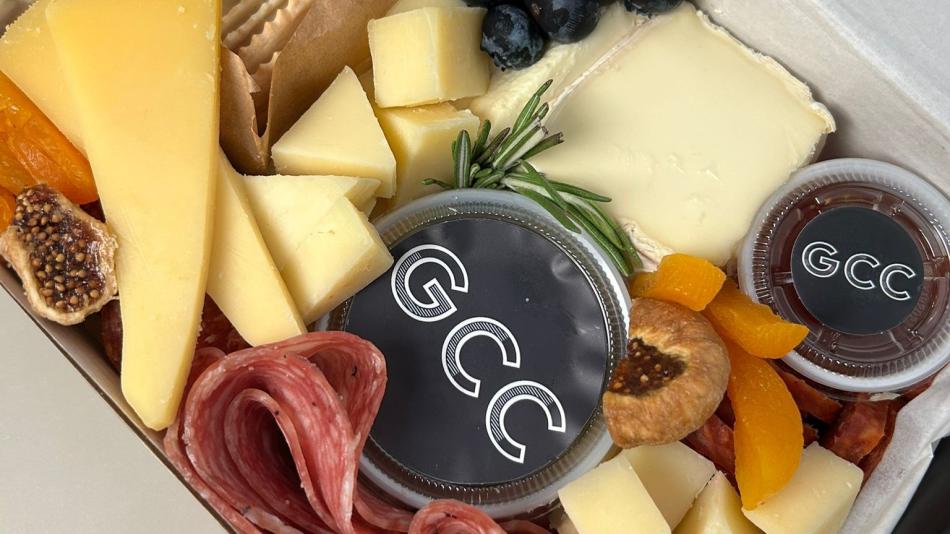 The image displays a close-up, top-down view of a charcuterie box, presented in a white cardboard container with dark gray accents. The box is densely packed with a variety of food items.