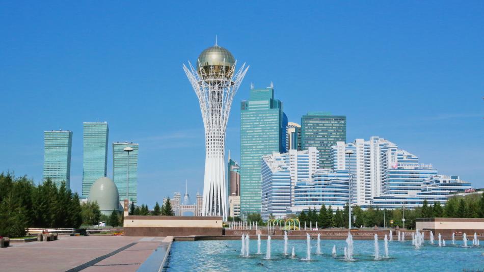 The image depicts a modern cityscape under a clear, bright blue sky. In the foreground, a wide, shallow fountain with multiple jets of water spraying upwards dominates the lower part of the frame. The water is a vibrant turquoise color. The area surrounding the fountain is paved with large, reddish-brown tiles interspersed with dark grey strips, forming a patterned walkway.