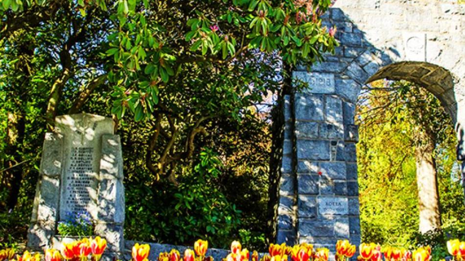 A stone arch and plaque sit at the edge of a forest surrounded by tulips.