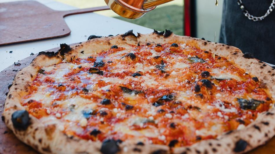 A close-up, eye-level shot captures a chef drizzling olive oil onto a freshly baked pizza. The pizza is the central focus, resting on a wooden peel, its crust puffed and speckled with dark, charred blisters, indicative of high-heat cooking. The topping is a vibrant combination of red tomato sauce and melted white mozzarella cheese, interspersed with fresh green basil leaves.