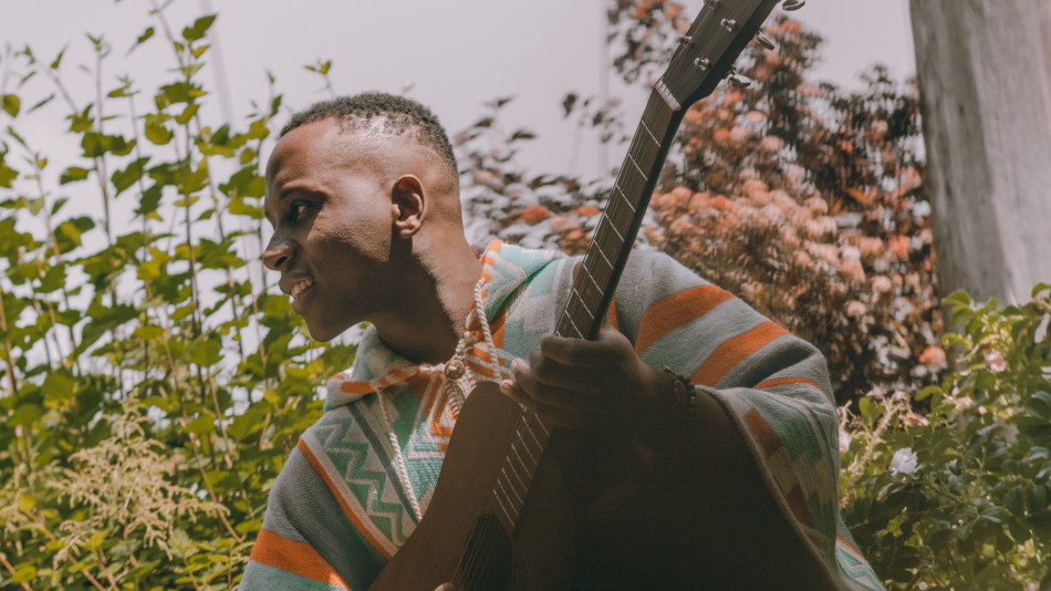 The image features a close-up, profile view of a Black man holding a guitar. He is wearing a poncho with a geometric pattern in shades of teal, orange, and grey. His expression is one of concentration or perhaps enjoyment, with his mouth slightly open and a smile visible. His skin tone is dark. He has short, textured hair.