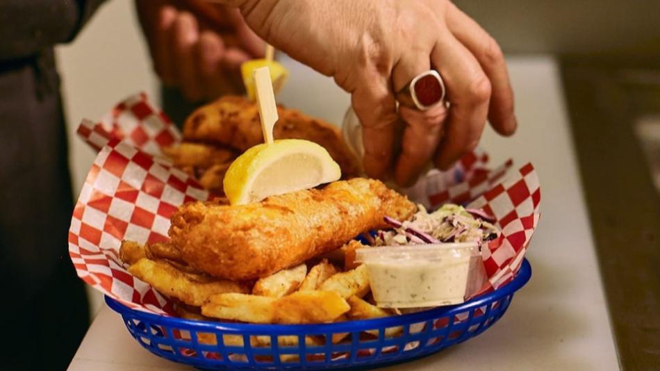 The image is a close-up, eye-level shot of a plate of fish and chips being prepared or served. The main focus is a blue plastic basket lined with red and white checkered paper. Inside the basket, there are several pieces of golden-brown, battered fish, and a generous portion of thick-cut french fries. A wedge of bright yellow lemon is perched on top of one of the fish pieces, secured with a wooden skewer. To the right of the fish and fries, a small clear plastic container holds a creamy white dipping sauce,