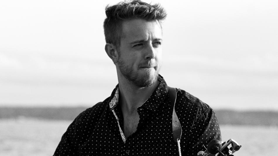 This is a medium shot, black and white photograph of a man holding a guitar. He is positioned slightly off-center to the left, with his head turned to the right, looking away from the camera. The man has light-colored hair styled upwards and a short, well-maintained beard. He is wearing a dark shirt with a repeating pattern of small white triangles or dots. The collar of the shirt is visible and has a contrasting white, patterned fabric. A dark leather strap from the guitar is visible across his chest.
