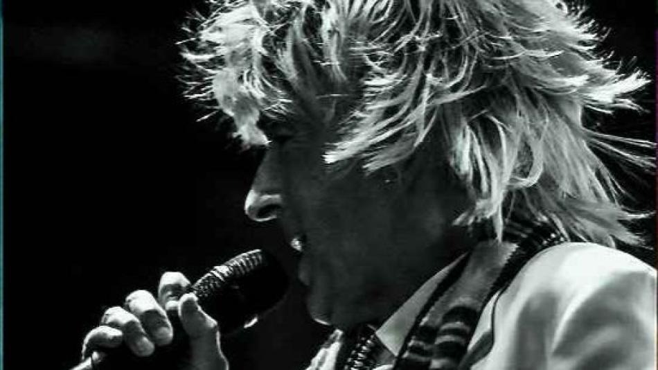 This is a black and white, close-up, profile shot of a male singer performing on stage. He has a full head of wild, spiky, blonde hair that frames his face and extends outward, suggesting movement. His head is tilted slightly downwards and to the left as he sings into a microphone held in his right hand. The microphone is dark and has a ribbed texture on its head. His mouth is open, and his eyes are not visible due to the angle.