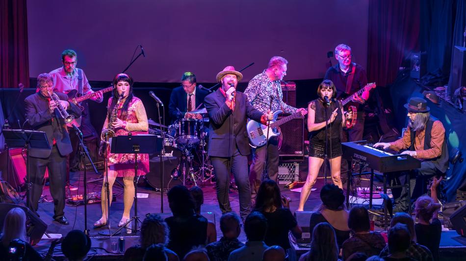 This is a medium shot of a live band performing on stage, captured under dramatic purple and blue stage lighting. The band consists of at least eight members, all actively engaged in their musical performance.