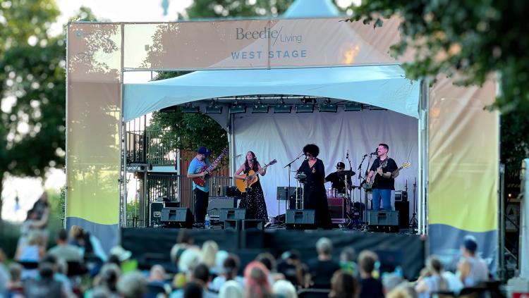 The image captures a vibrant outdoor concert on the Beedie Living West Stage, where a six-member band performs under a white canopy framed by peach and yellow banners. The musicians, playing guitars, drums, and singing, are surrounded by lush trees, adding to the relaxed summer atmosphere. A diverse audience sits on lawn chairs and blankets, enjoying the live music in what appears to be a community arts or music festival. The scene is lively yet laid-back, with golden hour lighting enhancing the festive, we