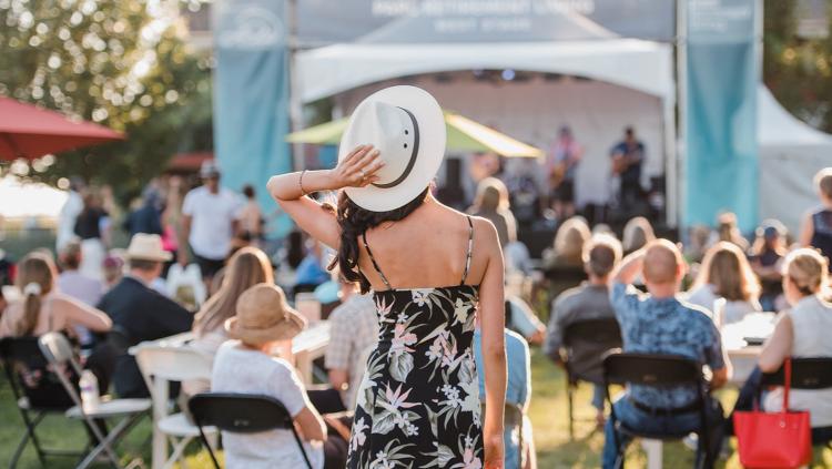 The image captures an outdoor event, likely a festival or concert, bathed in the warm glow of late afternoon or early evening sunlight. The scene is viewed from behind a woman in the foreground, who is the central subject. She has her back to the camera, wearing a black, spaghetti-strap dress with a bold floral pattern of pink and pale green flowers. She is also wearing a wide-brimmed white hat, tilted back slightly, with her right hand resting on its brim. A ring is visible on her right hand. Her dark hair