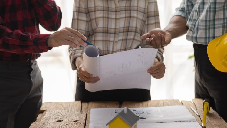 Three people stand over architectural plans and a model home on top of some paperwork.