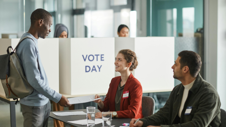 Two people wearing nametags sit at a table. One person is handing papers to someone standing in front of them. A sign in the background reads "Vote Day."