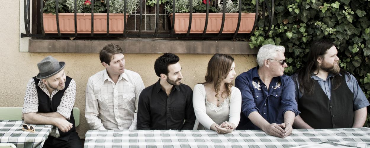 The image displays a group of six individuals, three men and three women, seated in a row at an outdoor table, likely a cafe or restaurant patio. They are all looking to their right, creating a unified gaze that draws the viewer's attention in that direction. The scene is set against a neutral beige wall and a window with decorative bars above it. Two terra-cotta planters filled with greenery and a few red flowers are positioned on a ledge beneath the window. To the right of the window, a dense wall of lush