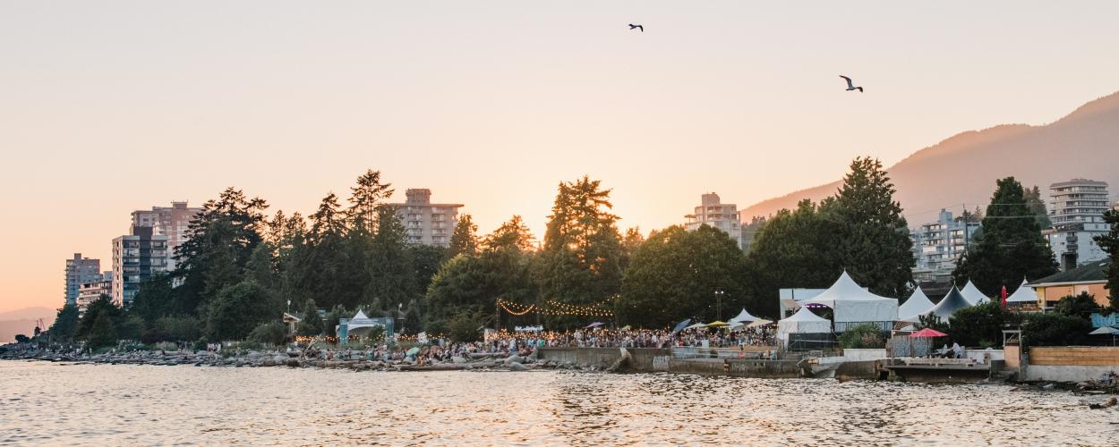 A large crowd gathers on the waterfront, enjoying a summer evening under the setting sun.  The scene is framed by a row of apartment buildings and lush green trees.  The sun casts a warm glow over the water, creating a sense of peace and tranquility.  A boardwalk extends into the water, and several white tents are set up on the shore, suggesting a festival or event.  The scene is alive with laughter and conversation, creating a vibrant and festive atmosphere. 