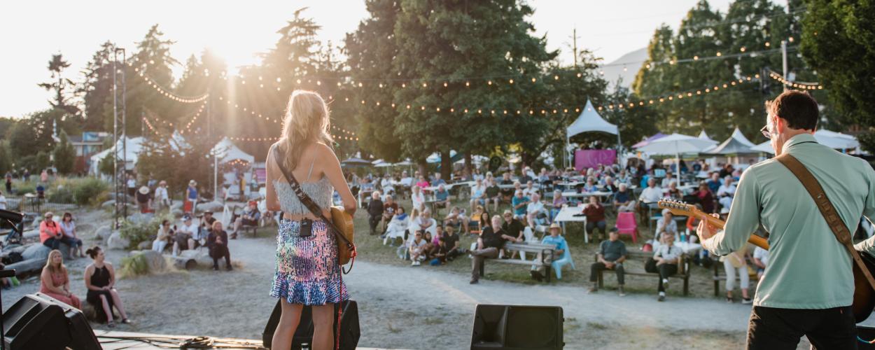 A live music performance takes place outdoors, illuminated by the warm glow of the setting sun. Two musicians, a woman with long blond hair and a man with dark hair, stand on a small stage, facing a crowd of people seated on the ground and benches. The woman is dressed in a sequined skirt and a white tank top, while the man wears a light green shirt and black pants. They both hold guitars, and the man is in the middle of playing his. The stage is equipped with two large speakers and a few microphones. The a