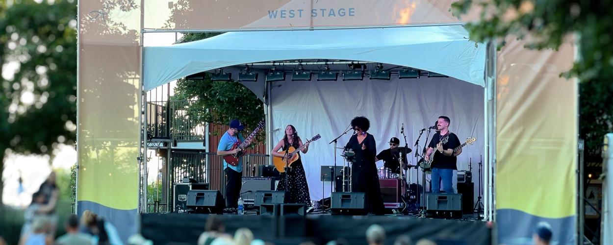 The image captures a vibrant outdoor concert on the Beedie Living West Stage, where a six-member band performs under a white canopy framed by peach and yellow banners. The musicians, playing guitars, drums, and singing, are surrounded by lush trees, adding to the relaxed summer atmosphere. A diverse audience sits on lawn chairs and blankets, enjoying the live music in what appears to be a community arts or music festival. The scene is lively yet laid-back, with golden hour lighting enhancing the festive, we