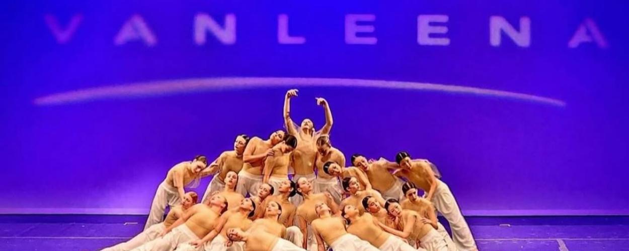 Full shot of a large group of dancers, likely a dance troupe, performing a contemporary dance.   The dancers are arranged in a complex, layered formation, with some dancers positioned atop others, forming a sort of pyramid or stacked arrangement. 