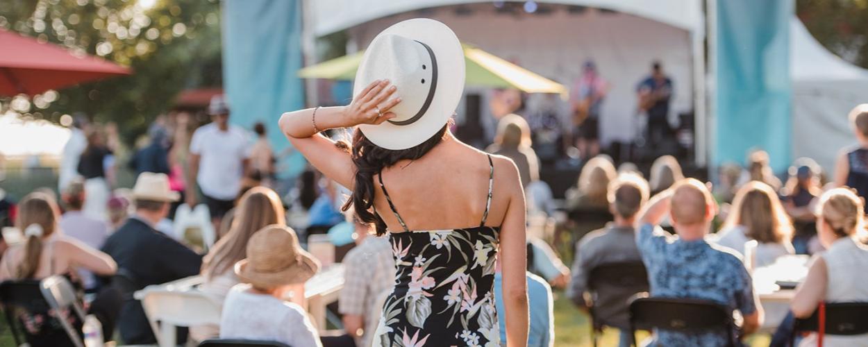 The image captures an outdoor event, likely a festival or concert, bathed in the warm glow of late afternoon or early evening sunlight. The scene is viewed from behind a woman in the foreground, who is the central subject. She has her back to the camera, wearing a black, spaghetti-strap dress with a bold floral pattern of pink and pale green flowers. She is also wearing a wide-brimmed white hat, tilted back slightly, with her right hand resting on its brim. A ring is visible on her right hand. Her dark hair