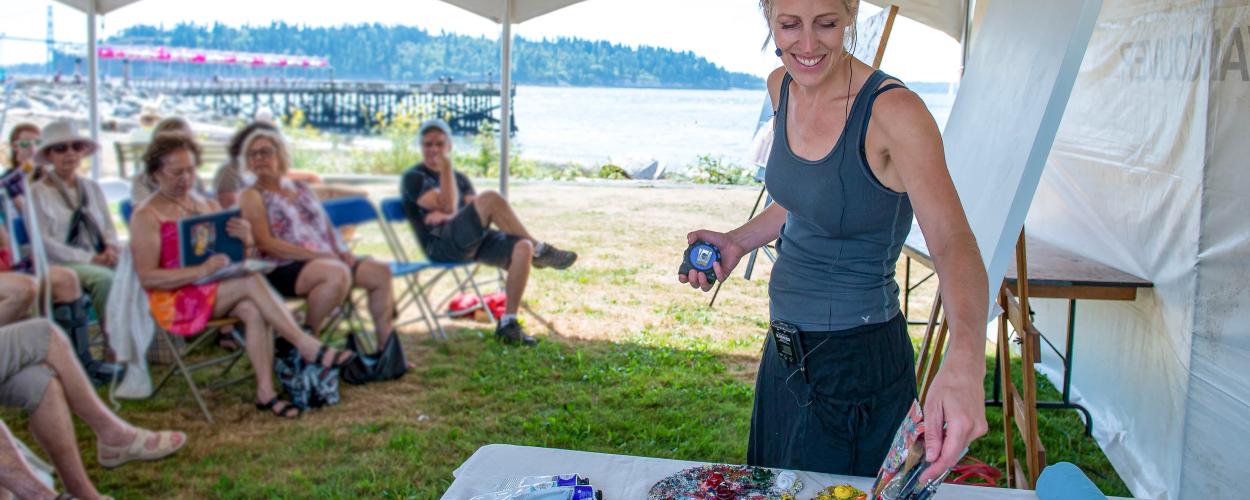 A woman is standing next to a table filled with art supplies. The setting appears to be outdoors, possibly during summer, surrounded by grass and a picnic atmosphere. There are also other people present in the scene, including a man and a group engaged in activities around a tent.