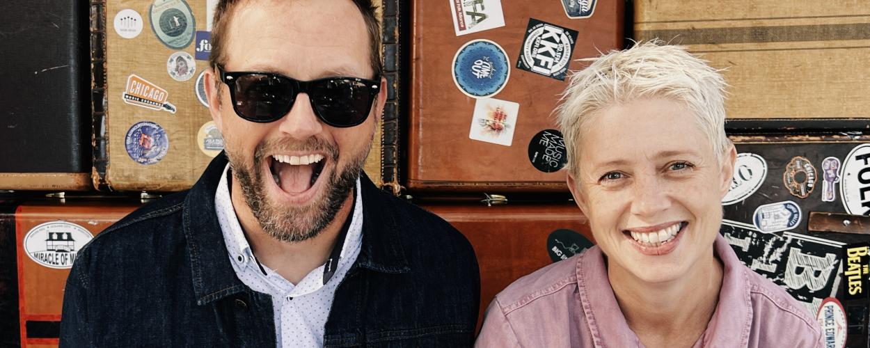 The image is a medium shot of a man and a woman, both smiling broadly and looking at the camera. They are positioned side-by-side in front of a wall of vintage suitcases.